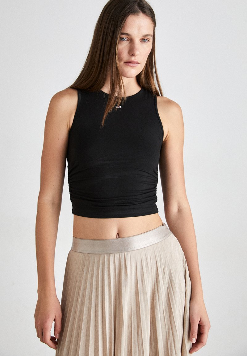 Top corto sin mangas negro con textura fruncida, combinado con una falda beige plisada que presenta una cinturilla brillante. Diseño simple y colores neutros.