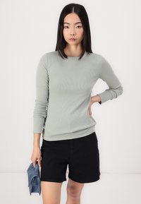 Lichtgroene langarm ribbeltop gecombineerd met zwarte shorts. De top heeft een ronde halslijn en een aangesloten ontwerp. Een blauwe clutch wordt in één hand vastgehouden.