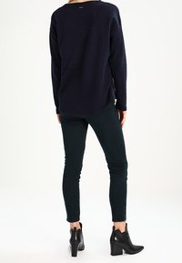 Persona de pie, de espaldas, vistiendo una camiseta de manga larga azul marino, pantalones oscuros ajustados y botas de tacón negras hasta el tobillo, sobre un fondo blanco.