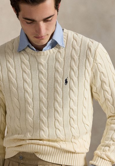 Homme portant un pull torsadé crème par-dessus une chemise bleu clair à col avec un pantalon beige, regardant vers le bas sur un fond neutre.