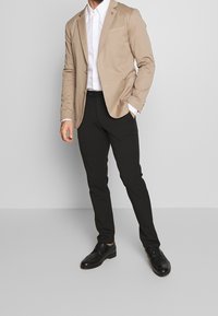 Beige bomullsblazer med en ficka, vit skjorta, svarta slim-fit byxor och svarta läderskor.