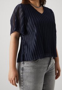 Marineblauw geplooid blouse met korte mouwen, lichte stof, V-hals en een ontspannen pasvorm. Gecombineerd met lichtgrijze denim voor contrast.