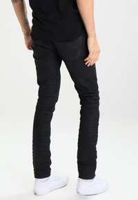 Osoba v černých skinny jeans a bílých teniskách, stojící s jednou rukou uvolněně vedle těla na světlém jednobarevném pozadí.