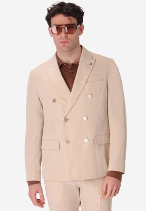 Uomo che indossa un blazer beige doppiopetto e pantaloni abbinati con camicia marrone sotto e occhiali da sole oversize marroni.