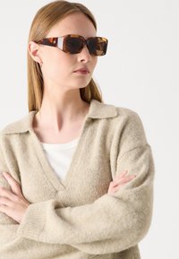 Brune skildpadde-solbriller med en tyk, vinkelret stel; båret med en lys beige, tekstureret striktrøje og en hvid top underneath.