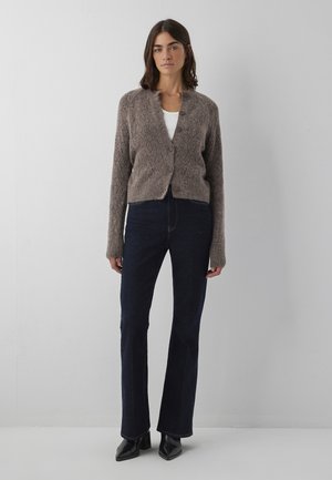 Frau steht vor einer weißen Wand und trägt eine braune, geknöpfte Strickjacke, ein weißes Hemd, dunkelblaue Jeans und schwarze Stiefel mit Absatz.