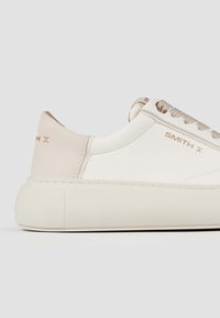 Sneaker bianca in pelle con accenti rosa pallido, suola in gomma spessa, occhielli metallici e marchio in rilievo "SMITH X" sul lato e sul tallone.