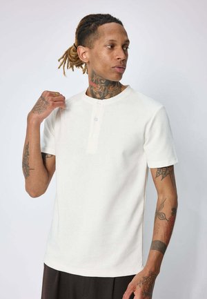 Homme avec des tatouages et des dreadlocks portant un t-shirt henley blanc à manches courtes regardant sur le côté sur un fond clair uni.