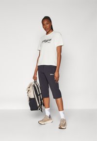 Lichtgekleurde t-shirt met graphic, donkere cropped broek, beige sneakers, witte sokken. Houdt een beige en zwarte rugzak vast.