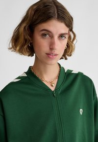 Nuori nainen, jolla on lyhyet aaltoilevat hiukset, pukeutuneena vihreään vetoketjulliseen toppiin, jossa on valkoiset raidat, ja kullanväriseen sydämen muotoiseen riipukseen kaulaketju yksivärisellä taustalla.