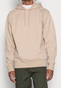 Sudadera beige con capucha hecha de tela suave, que cuenta con un bolsillo canguro, capucha con cordón y puños y dobladillo acanalados.