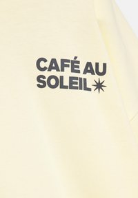 Material galben deschis cu text negru îngroșat „CAFÉ AU SOLEIL” și un simbol negru de stea cu opt colțuri în partea dreaptă.