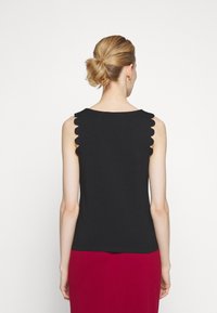 Femme de dos, cheveux relevés en un chignon soigné, portant un haut noir sans manches à bords festonnés et une jupe rouge, debout devant un fond uni.