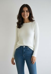 Kobieta o długich ciemnych włosach, ubrana w biały sweter i niebieskie dżinsy, stojąca i uśmiechająca się na jasnym, jednolitym tle.