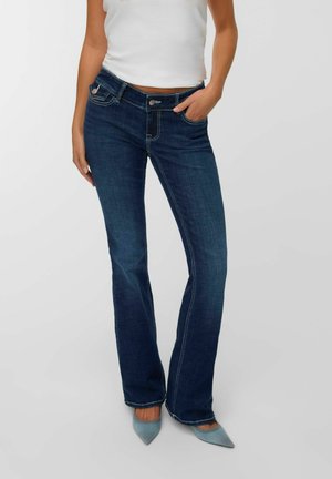 Kvinde iført mørkeblå bootcut jeans med en mellemhøj talje, hvid udenærmet top og lyseblå spidse sko.