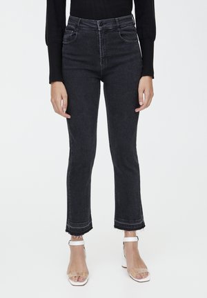 Woman wearing high-waist black cropped jeans with frayed hems and white open-toe ankle-strap heels, standing against a white background.