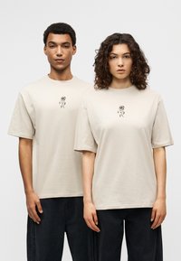Μπεζ βαμβακερά t-shirts με κοντά μανίκια, στολισμένα με μαύρο κεντητό λουλούδι στο στήθος, συνδυασμένα με σκούρα τζιν παντελόνια.