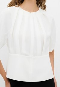 Femme portant un chemisier blanc avec des détails plissés, des manches courtes bouffantes et un col rond, associé à un pantalon noir.