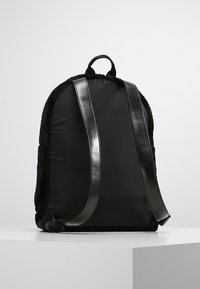 Sac à dos noir en matériau doux et pelucheux avec des sangles en cuir lisse et une forme arrondie. Comprend une poignée supérieure pour le transport.
