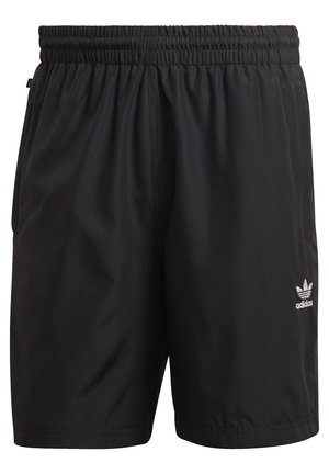 Mężczyzna w ciemnozielonym t-shircie Adidas, czarnych szortach Adidas i białych adidasach z czarnymi paskami, stoi z rękami w kieszeniach.