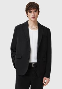 Schwarzer Blazer mit Reverskragen, zwei Knöpfen und Fronttaschen. Über einem weißen gerippten Hemd getragen. Minimalistisches Design mit taillierter Passform.