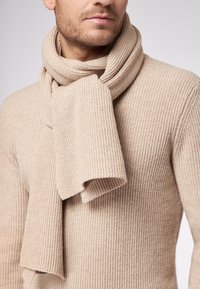 Mand iført en beige ribstrikket sweater og et matchende tørklæde viklet om halsen foran en ensfarvet baggrund.