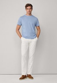 Mies, joka seisoo yksinkertaista seinää vasten, päällä vaaleansininen lyhythihainen paita, valkoiset housut ja ruskeat mokkanahkaiset loaferit, kädet taskuissa.