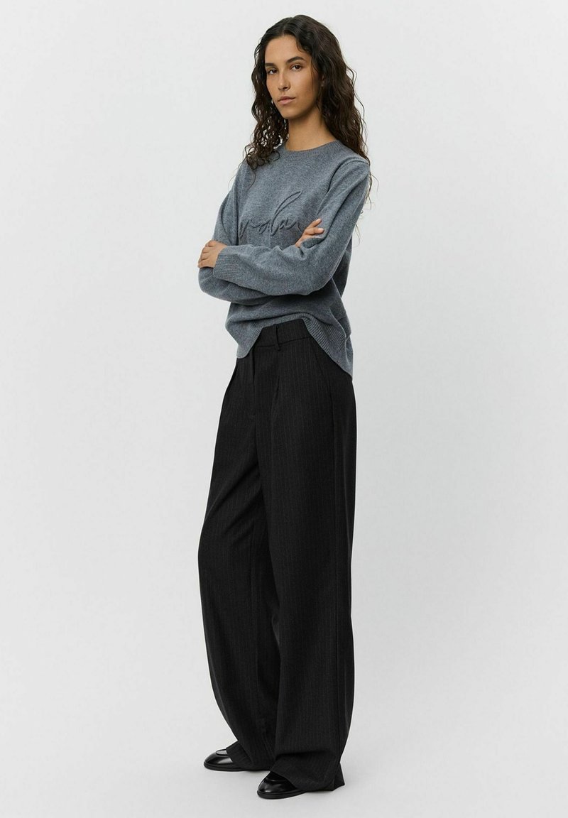 Maglione grigio in maglia con maniche lunghe e dettagli sottili, abbinato a pantaloni larghi a righe verticali neri. Tessuto morbido e vestibilità rilassata.