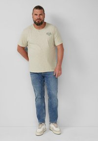 Helles beige T-Shirt mit kurzen Ärmeln und rundem Ausschnitt, verziert mit grünem Grafikdruck, kombiniert mit blauen Jeans und weißen Sneakern.