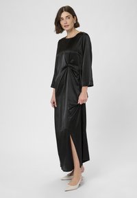 Frau, die ein langes schwarzes Satin-Kleid mit seitlichem Schlitz und gerafftem Detail an der Taille trägt, kombiniert mit weißen spitzen Slingback-Absätzen.