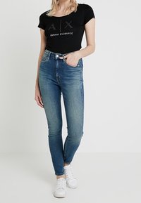 Černé tričko s krátkým rukávem s logem "A/X Armani Exchange", doplněné středně vysokými modrými skinny džíny a bílými teniskami. Hladká džínová textura.