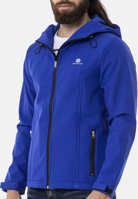 Blaue Softshelljacke mit Kapuze, Frontreißverschluss und Seitentaschen. Hat schwarze Akzente und ein Logo auf der linken Brust.