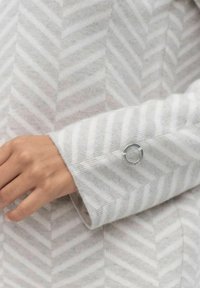 Manche de pull gris rayé avec un motif chevron, texture douce et un accent circulaire en argent sur le poignet.