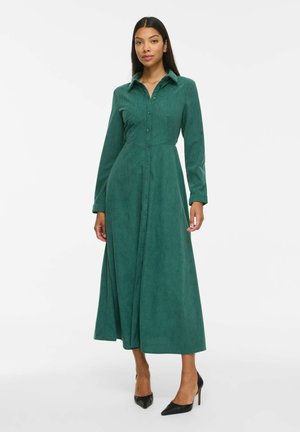 Femme porte une robe midi verte à manches longues, boutonnée, avec col, ainsi que des chaussures à talons noirs, debout devant un fond blanc uni.