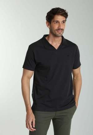 Homme portant un polo noir à manches courtes et un pantalon vert olive, debout avec une main dans la poche, regardant légèrement vers la droite.