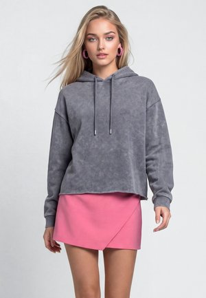 Jonge vrouw met lang blond haar, die een grijze hoodie, een roze wikkelrok en roze creolen draagt, staand tegen een effen achtergrond.