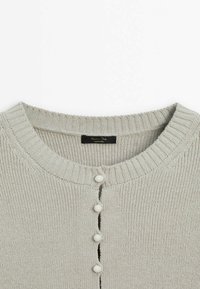 Vaaleanharmaa neulottu neuletakki pyöreällä pääntiellä ja viisi valkoista nappia edessä, näkyvä merkkilappu kauluksen sisäpuolella.