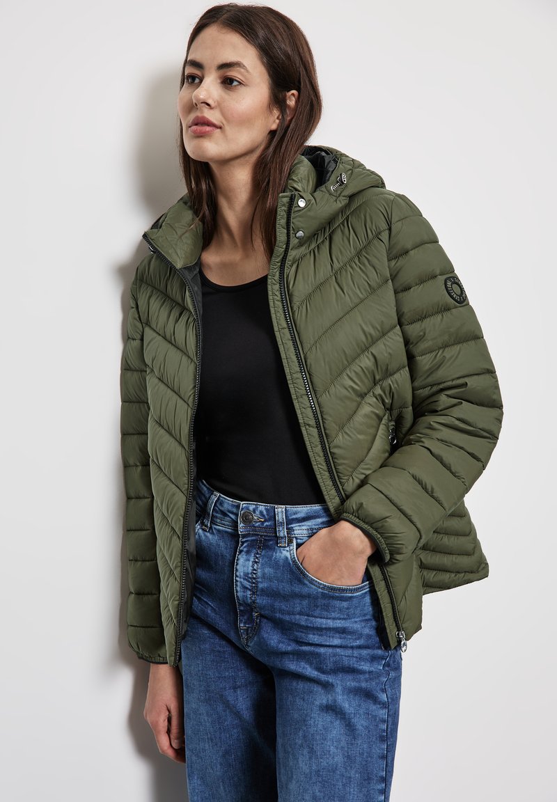 Doudoune vert olive avec fermeture à glissière et pression, dotée d'une capuche et d'un motif matelassé horizontal, associée à un jean en denim bleu.