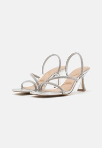 Sølvbelagte sandaler med flere tynde stropper, der har en skinnende metallisk finish og et unikt, vinklet hældesign. Beige sål.
