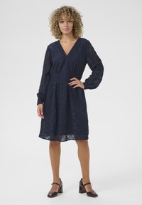 Abito in pizzo blu navy con scollatura a V, maniche lunghe e vita cintrata, caratterizzato da un motivo floreale e una gonna a campana. Abbinato a tacchi marroni.