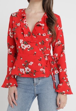 Femme portant un chemisier cache-cœur à fleurs rouge avec un col à volants et des manches évasées, associé à un jean bleu clair.
