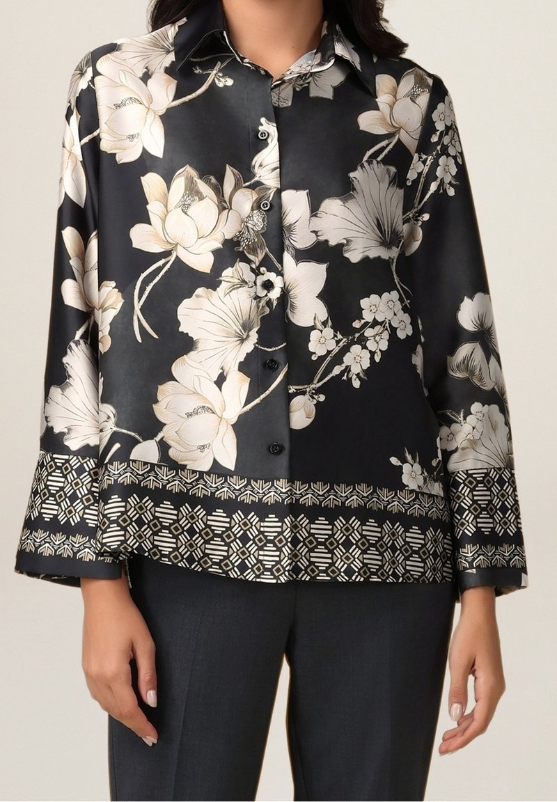 Blouse noire à manches longues boutonnée avec grand imprimé floral blanc et bordure à motif géométrique, portée avec un pantalon foncé.