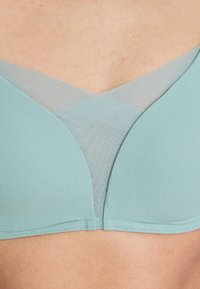 Light teal bra with soft mesh fabric detailing forming a crisscross pattern at the center front on bare skin background.