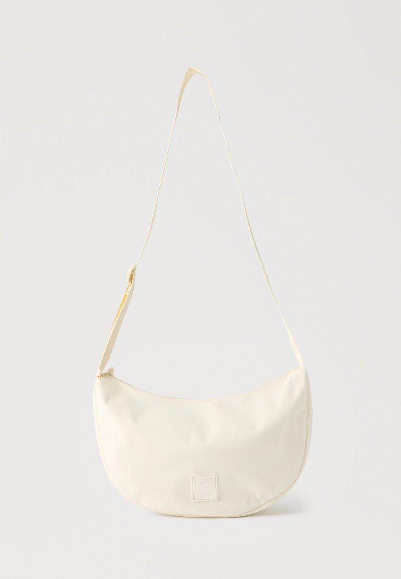 Σακίδιο ώμου σε κρεμ απόχρωση σε σχήμα ημισελήνου με ρυθμιζόμενο λουράκι και μικρό λογότυπο μπροστά, σε απλό φόντο.