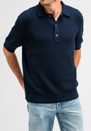 Navy blue knitted polo shirt with ribbed texture, short sleeves, three-button placket, and fitted hem. Paired with light denim jeans.