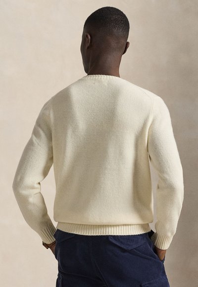 Man wearing a plain cream-colored knit sweater and dark blue pants, standing with hands in pockets, seen from the back against a beige background.