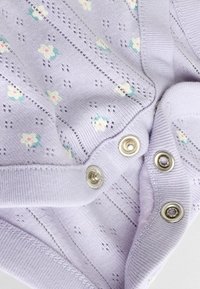 Lawendowa dzianina z małymi, białymi i turkusowymi haftowanymi kwiatami oraz srebrnymi zatrzaskami, ukazująca szczegóły fakturowanego szycia.