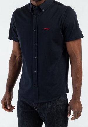 Homme portant une chemise noire à manches courtes avec un petit logo rouge sur la poitrine et un jean foncé sur un fond uni.