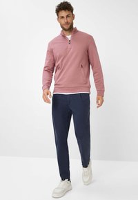 Felpa rosa con zip, dotata di due tasche laterali, abbinata a pantaloni blu e sneaker bianche. Tessuto morbido, vestibilità casual e design moderno.