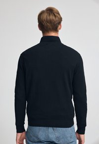 Suéter negro de punto con un cuello de canalé, mangas largas y un dobladillo ajustado, mostrado desde la parte de atrás con una textura suave.
